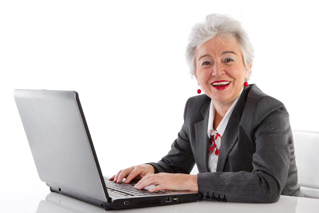 Ältere Hauswirtschaftsmeisterin sitzt am Schreibtisch mit einem Laptop und lächelt in die Kamera
